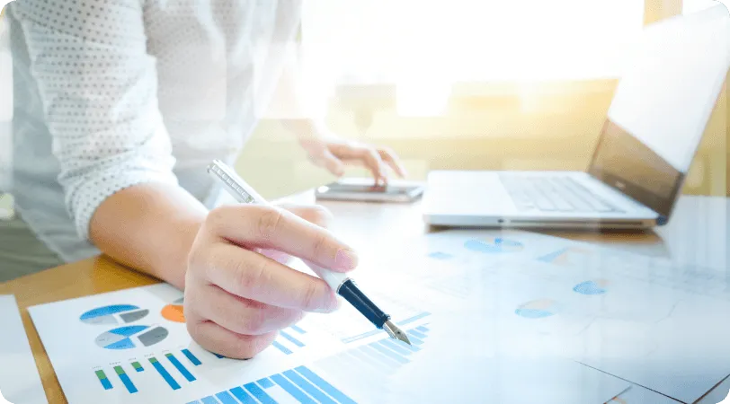 Hand holding a pen over financial charts next to a laptop